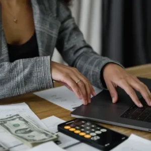 A business professional using a laptop and calculator surrounded by payslips, cash, and financial documents.