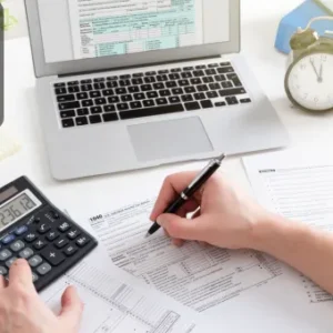 A person using a calculator and completing financial documents, with a laptop displaying accounting software in the background.