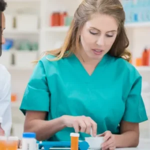 A healthcare worker in scrubs carefully preparing medication under supervision in a pharmacy setting.