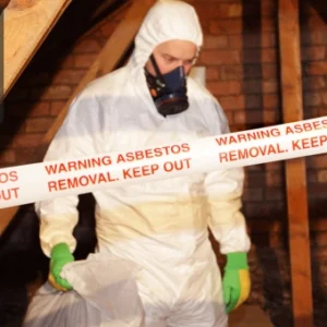 Worker in protective gear handling asbestos material inside a sealed-off area marked with warning tape.