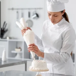 A pastry chef in a white uniform decorating a cake with cream using a piping bag in a professional kitchen.