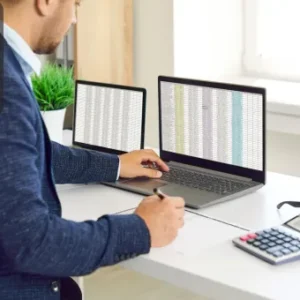 An office administrator working on a laptop with spreadsheets displayed on the screen, using a pen and calculator at a tidy desk.