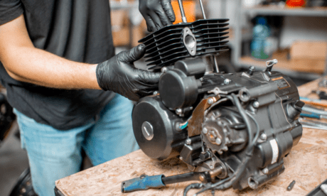 Mechanic in overalls working on a motorcycle engine in a garage, with tools and a dismantled bike engine on a workbench.