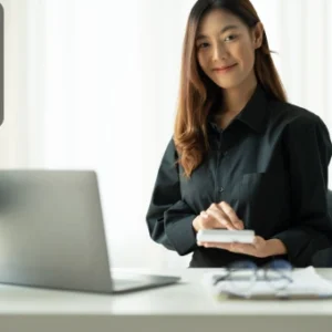 Professional woman in a black shirt working on a laptop and holding a phone, symbolising productivity and organisation in the workplace.