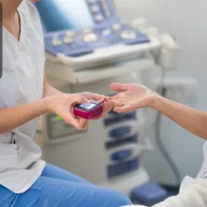 A healthcare professional performs a blood glucose test on an elderly patient using a handheld monitor in a clinical setting. Thames College logo is displayed in the top-left corner.