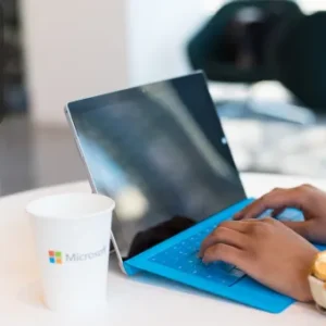 A person typing on a Microsoft Surface tablet with a blue keyboard cover, seated at a white table with a Microsoft-branded cup. The Thames College logo is visible in the top left corner.