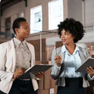 Two professional women in a warehouse discussing operations with digital tablets in hand.