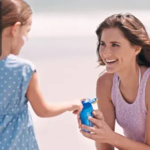 A smiling woman kneels on a beach holding sunscreen while speaking with a young girl in a blue dress; Thames College logo is displayed in the top-left corner.