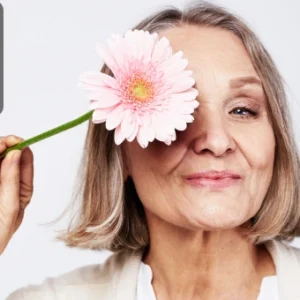 An elderly woman smiling while holding a flower over one eye, symbolising positivity and vitality in later life.