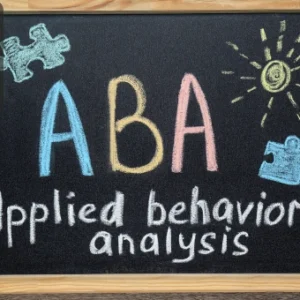 A chalkboard with the letters “ABA” in colourful chalk and the words “Applied behavior analysis” written below, decorated with puzzle pieces and a sun drawing.
