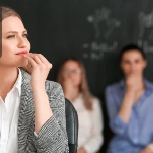 A woman learning British Sign Language in a classroom, practising hand signs with others in the background.