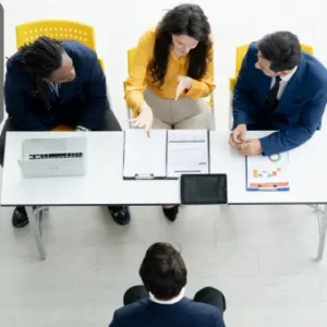 A top-down view of a job interview panel with three interviewers across a desk from a candidate, reviewing documents and engaging in discussion; Thames College logo appears in the top-left corner.
