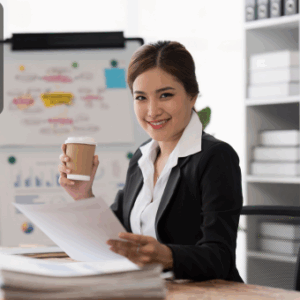 Professional woman in formal attire smiling while holding financial documents and a coffee cup at her desk, with charts and files in the background, symbolising expertise in accounting and payroll.