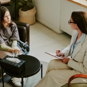 A professional counsellor holding a clipboard speaks empathetically with a young woman during a counselling session in a calm, comfortable office. The Thames College logo is visible in the corner.