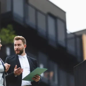 Two business professionals discussing property investment details outdoors near modern buildings, holding documents and engaged in conversation.