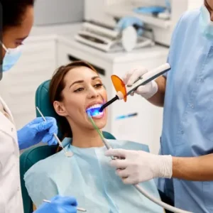 A dental assistant supports a dentist during a procedure while a patient receives treatment under a curing light.