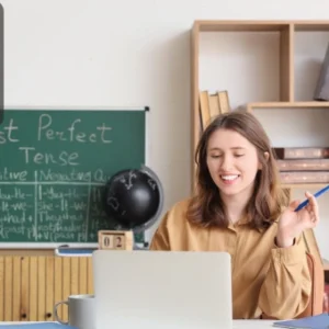 Smiling teacher sitting at a desk with a laptop, bookshelves behind her, and a blackboard showing grammar notes, representing a blended learning course.