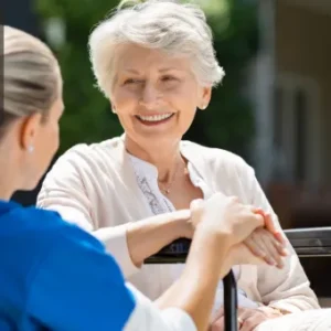 A smiling elderly woman sits outdoors in a wheelchair while a female caregiver in uniform gently holds her hands and talks to her. The scene radiates warmth, care, and trust. The Thames College logo appears in the top-left corner.
