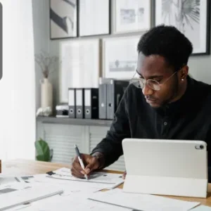 A focused professional writing notes at a desk with papers and a tablet, surrounded by office supplies and documents.