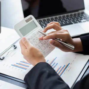 A professional using a calculator while analysing printed financial graphs, with a laptop in the background.
