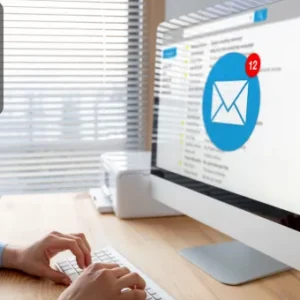 A person typing on a keyboard with a large desktop screen displaying an email inbox with new messages.