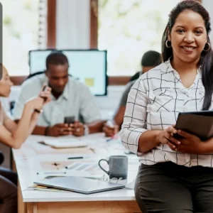 Confident businesswoman holding a tablet and smiling in an office, with a team collaborating in the background, symbolising entrepreneurship and business planning.
