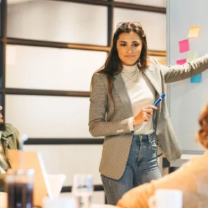 A woman leading a team meeting while pointing to sticky notes on a whiteboard, demonstrating Agile Scrum project management.
