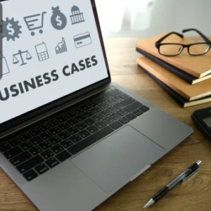 Laptop displaying the words “Business Cases” on the screen beside a calculator, notebooks, pen, and glasses on a wooden desk, with the Thames College logo in the corner.