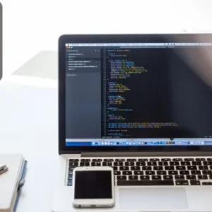 A laptop on a white desk displaying Python code in a text editor, with a smartphone, notebook, and pen nearby; Thames College logo is visible in the top corner.