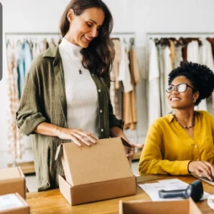 Two women working together in a clothing store, packing boxes and using a laptop to manage their business.