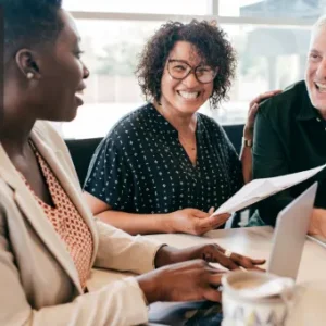 Group of professionals discussing business documents and smiling during a collaborative meeting with a laptop and coffee on the table.
