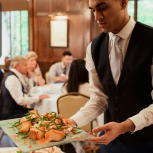 A catering staff member in formal uniform serves a plate of gourmet food to guests seated at a dining event. The Thames College logo is visible in the corner.