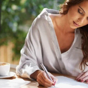 Woman writing in a notebook with coffee beside her, symbolising creativity and the journey to becoming an author.