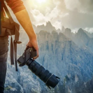 Photographer holding a professional camera while standing on a mountain with sunlight and rocky peaks in the background, featuring the Thames College logo in the corner.