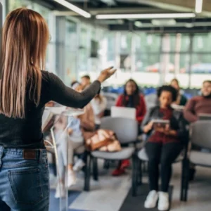 A female trainer addressing a small group of adult learners in a bright, modern classroom, representing motivation and professional growth, with the Thames College logo in the top-left corner.