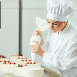 A pastry chef in uniform skillfully piping cream onto cakes in a professional bakery setting.