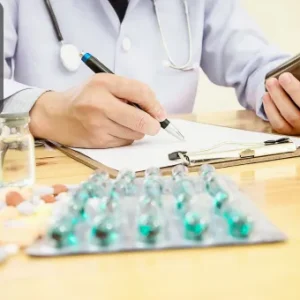 A healthcare professional writing on a clipboard while holding a medicine bottle, with tablets and ampoules on the table.