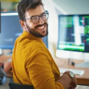 A smiling man wearing glasses and a mustard shirt sits in front of a desktop monitor displaying complex code, with another person working beside him; Thames College logo is shown in the top corner.