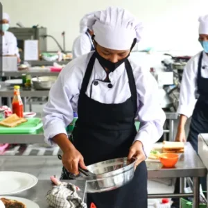 A group of chefs in professional uniforms and aprons work in a commercial kitchen, preparing and plating food with attention to hygiene. One chef wears a face mask. The Thames College logo is displayed in the top left corner.
