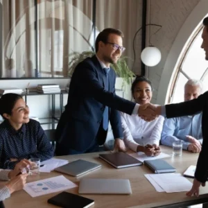 Business professionals in a meeting, with two men shaking hands while colleagues look on, symbolising HR management, compliance, and conflict resolution.