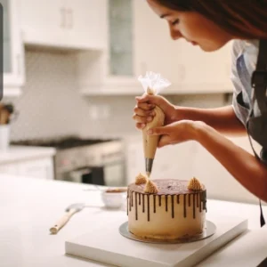 A baker carefully decorating a chocolate drip cake with piped cream swirls in a modern kitchen.