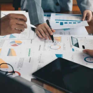 Team members analysing colourful charts and graphs on a wooden table, using pens, tablets, and printed business reports during a management and analysis meeting.