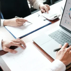 Two professionals reviewing financial documents with charts and graphs displayed on a laptop screen, along with notes and a calculator on the table.