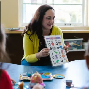 A teacher sits with young children, holding up a colourful phonics chart with letters and illustrations, engaging them in a phonics learning activity. The Thames College logo is visible in the top left corner.