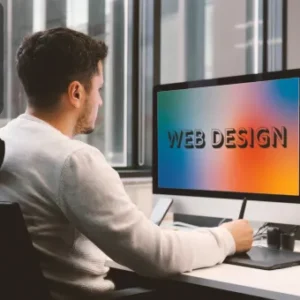 A man seated at a desk in a modern office setting is working on a computer displaying the words “WEB DESIGN” on a colourful gradient screen; Thames College logo is shown in the top corner.