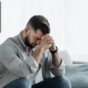 A distressed man sitting on a sofa with his head bowed and hands clasped in front of his face, showing visible signs of anxiety and emotional strain.