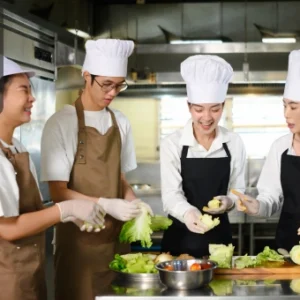 Group of culinary students in chef uniforms preparing vegetables together in a professional kitchen setting.
