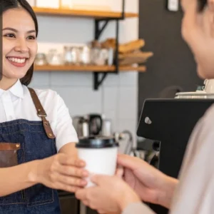 A smiling barista handing a takeaway coffee cup to a customer in a café.