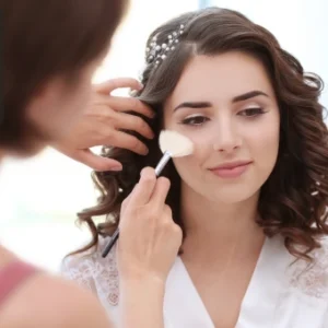 Makeup artist applying bridal makeup with a brush to a young woman with styled hair.