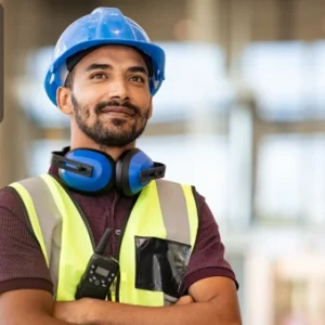 Confident construction worker wearing a safety helmet, reflective vest, and ear protection, standing on a building site with the Thames College logo in the corner.